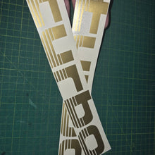 Load image into Gallery viewer, Turbo decal with gold and black design held over a green cutting mat.
