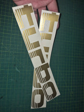 Load image into Gallery viewer, Turbo decal with gold and black design held over a green cutting mat.