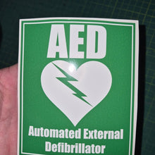 Load image into Gallery viewer, Green and white sticker with AED (Automated External Defibrillator) logo held by a hand on a dark grid background.