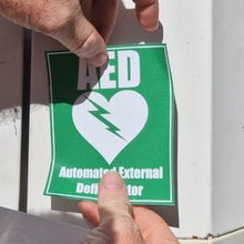 Load image into Gallery viewer, Person holding a green AED (Automated External Defibrillator) sticker with a white heart and lightning bolt symbol.