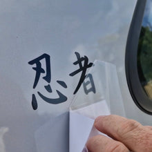 Load image into Gallery viewer, Hand applying a transparent decal with black text on a vehicle window.