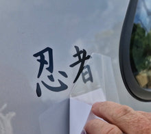 Load image into Gallery viewer, Hand applying a transparent decal with black text on a vehicle window.