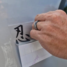 Load image into Gallery viewer, Hand with a ring writing a black character on a white board Black Japanese text saying ninja! Decal