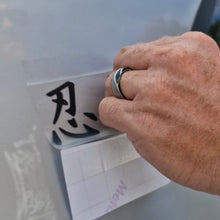 Load image into Gallery viewer, Hand with a ring writing a black character on a white board Black Japanese text saying ninja! Decal