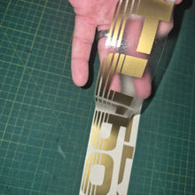 Load image into Gallery viewer, Gold and white decal held over a green cutting mat
