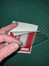 Load image into Gallery viewer, Hand holding a small book or card with a red cover against a green grid background