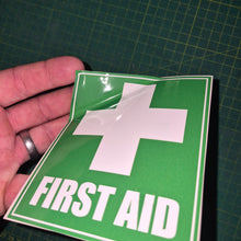 Load image into Gallery viewer, Green 'First Aid' sign with a white cross held by a hand on a grid background