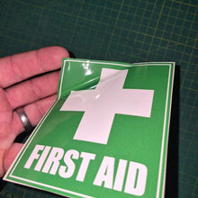 Load image into Gallery viewer, Green 'First Aid' sign with a white cross held by a hand on a grid background