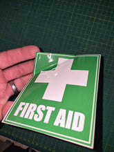 Load image into Gallery viewer, Green 'First Aid' sign with a white cross held by a hand on a grid background