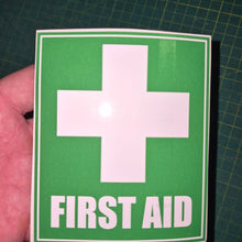 Load image into Gallery viewer, First aid sign with a cross on green paper held by a hand on a grid background