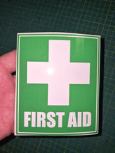 Load image into Gallery viewer, First aid sign with a cross on green paper held by a hand on a grid background