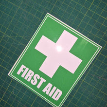 Load image into Gallery viewer, First aid sign with a white cross on a green background, placed on a grid surface.