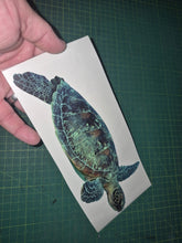 Load image into Gallery viewer, Hand holding a decal of a green sea turtle on a white card with a grid-patterned surface in the background.