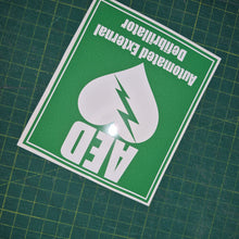 Load image into Gallery viewer, Green and white sticker with AED (Automated External Defibrillator) text and symbol on a grid background