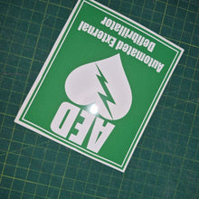 Load image into Gallery viewer, Green and white sticker with AED (Automated External Defibrillator) text and symbol on a grid background