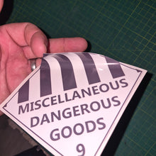 Load image into Gallery viewer, Hand holding a triangular label with 'Miscellaneous Dangerous Goods' text on a green cutting mat.