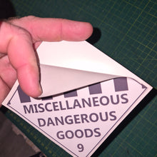 Load image into Gallery viewer, Hand holding a triangular label with 'Miscellaneous Dangerous Goods' text on a green cutting mat.