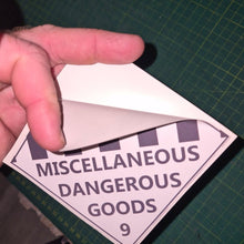 Load image into Gallery viewer, Hand holding a triangular label with 'Miscellaneous Dangerous Goods' text on a green cutting mat.