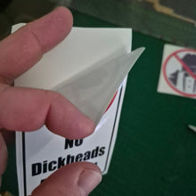 Load image into Gallery viewer, Hand holding a small white object over a 'No Dickheads' Sticker on a green surface.