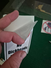 Load image into Gallery viewer, Hand holding a small white object over a 'No Dickheads' Sticker on a green surface.