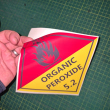 Load image into Gallery viewer, Organic Peroxide 5.2 Sticker Hand holding a red and yellow warning label on a green cutting mat