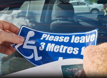 Load image into Gallery viewer, Hand holding a blue and white decal with a wheelchair icon and text 'Please leave 3 Metres' in front of a car.