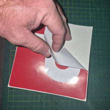 Load image into Gallery viewer, Hand peeling a white sticker from a red and white square on a green cutting mat.
