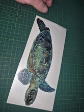 Load image into Gallery viewer, Sticker of a turtle on a piece of paper held by a hand on a green cutting mat.