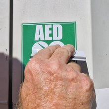 Load image into Gallery viewer, Hand pressing a button on an AED machine with a green sign above it.