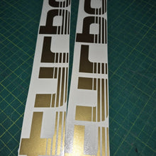Load image into Gallery viewer, Two gold and black car decals on a green cutting mat.