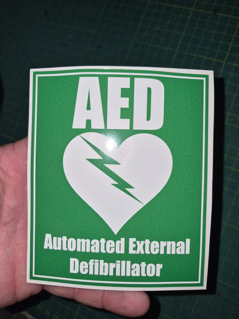 Green and white sticker with AED (Automated External Defibrillator) logo held by a hand on a dark grid background.