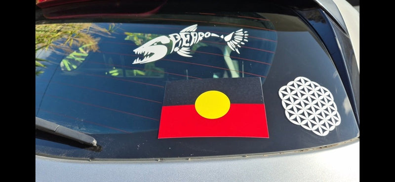 Car window with three stickers: an Aboriginal flag, a white geometric design, and a black and white animal emblem.