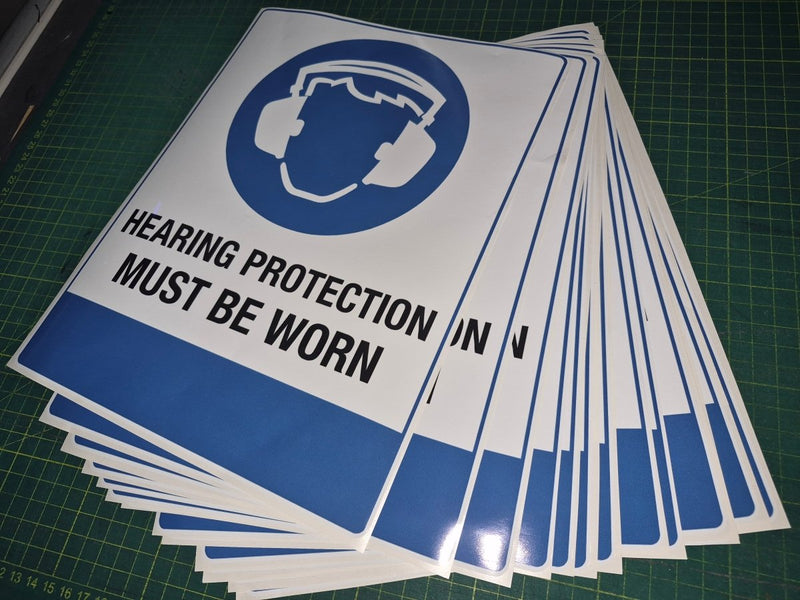 Stack of 'Hearing Protection Must Be Worn' signs on a green cutting mat.