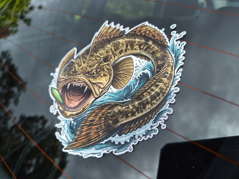 A die-cut vinyl sticker of a detailed flatty flathead fish jumping through blue water with a green lure in its mouth, shown applied to a dark tinted car window.