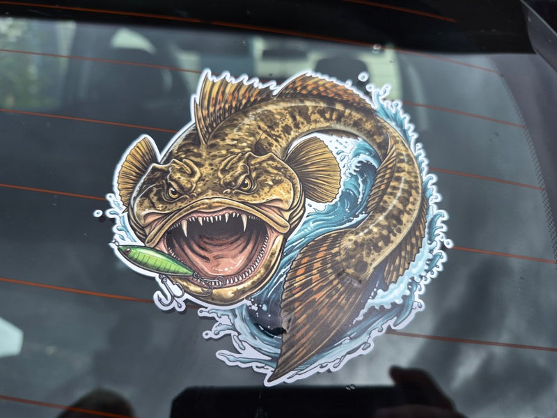 A die-cut vinyl sticker of a fierce, mouth-open flathead fish holding a green lure, applied to a dark car window.