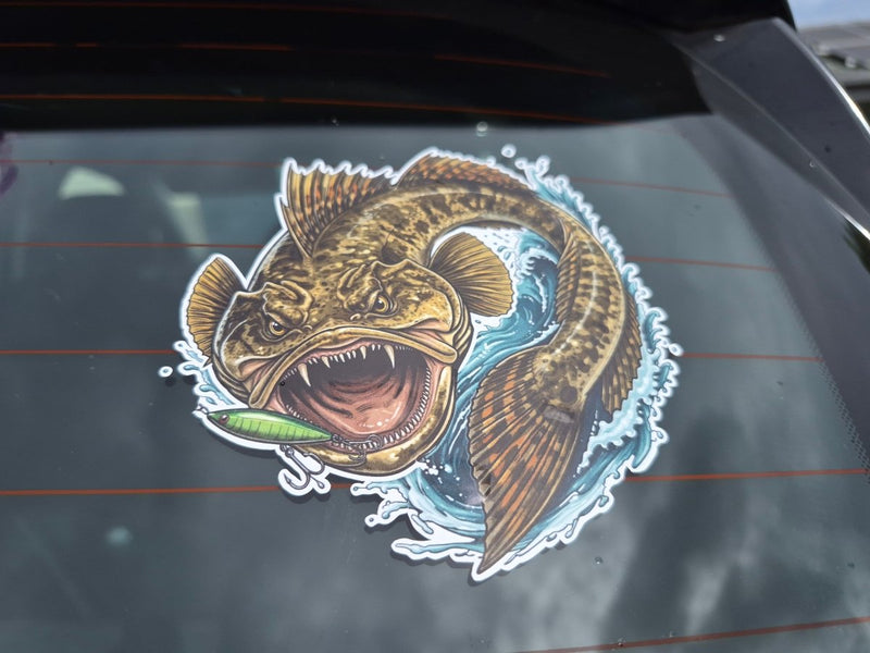 A die-cut vinyl sticker of a detailed fishing flatty fish holding a green lure, shown applied to the rear window of a vehicle.