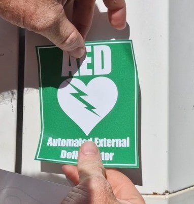 Person holding a green AED (Automated External Defibrillator) sticker with a white heart and lightning bolt symbol.