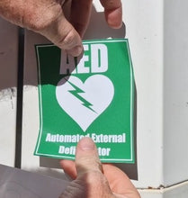 Load image into Gallery viewer, Person holding a green AED (Automated External Defibrillator) sticker with a white heart and lightning bolt symbol.
