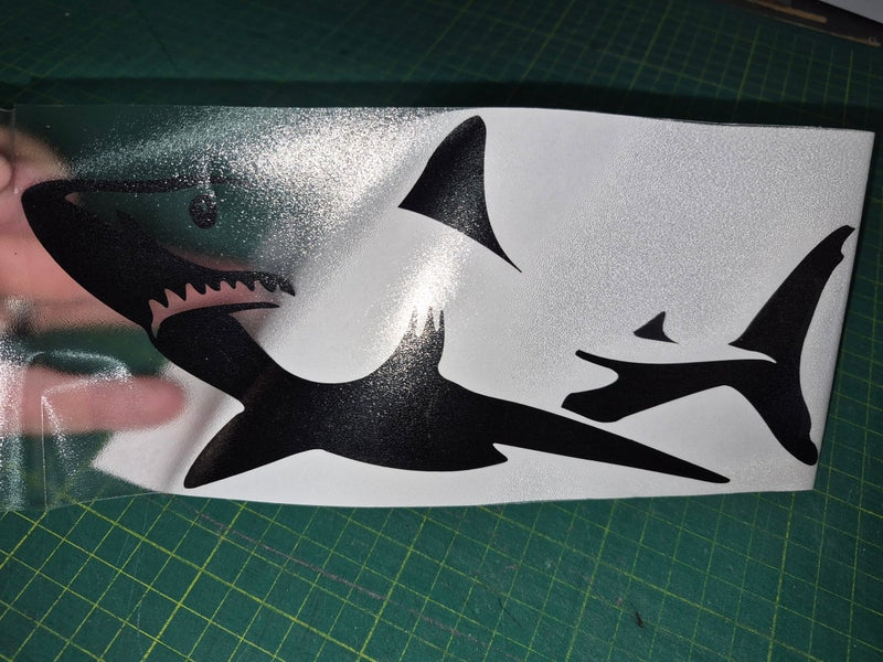 Black shark decal on a white sheet with a hand touching it, on a green cutting mat.