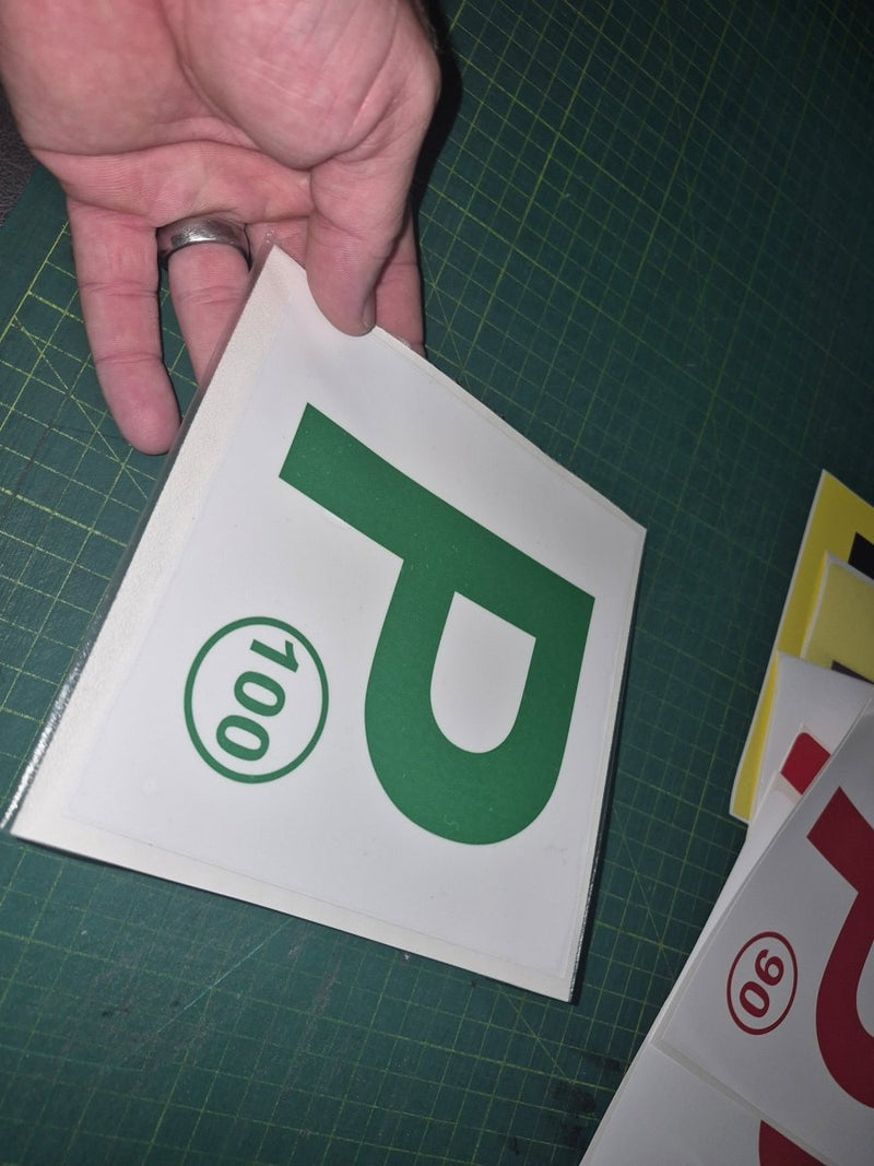 Hand holding a green 'P' with number 100 on a white background, on a green cutting mat.