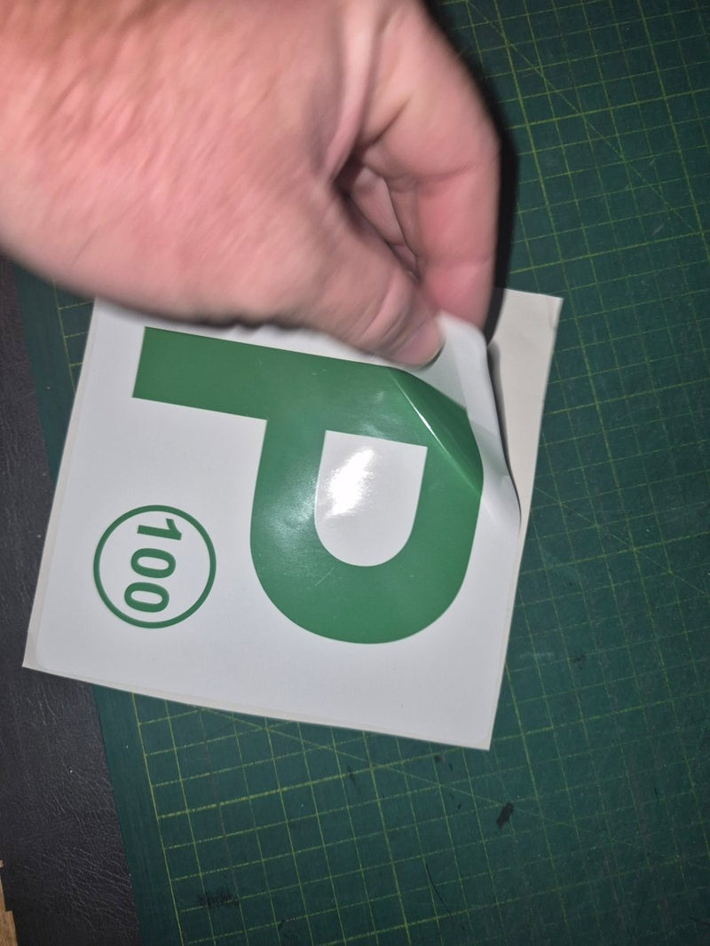 Hand holding a green and white 'P' decal with the number 100 on a green cutting mat.
