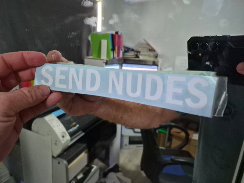 Hand holding a sticker with 'SEND NUDES' text in an indoor setting.