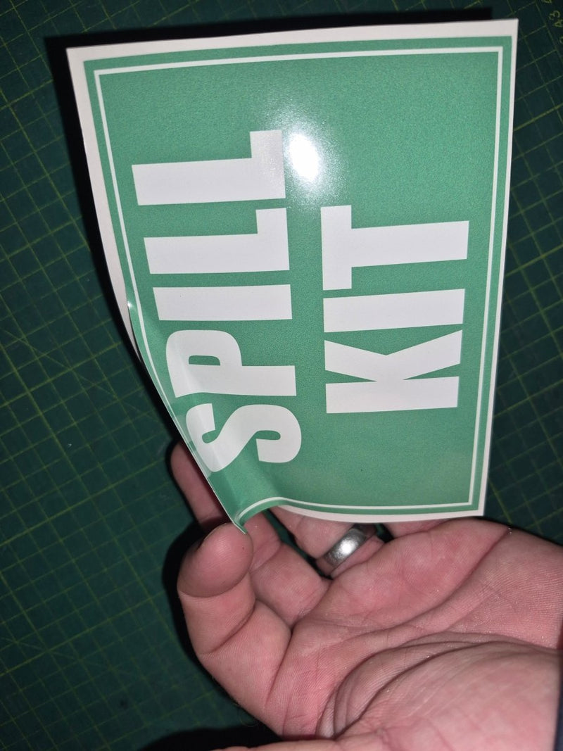 Hand holding a green sign with 'SPILL KIT' text on a cutting mat background