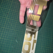 Load image into Gallery viewer, Gold and white decal held over a green cutting mat
