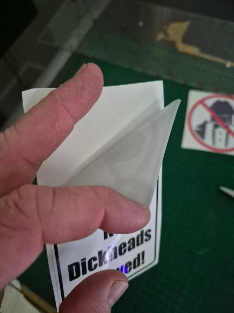 Hand holding a small white object with a green surface and 'Dickhead' sticker in the background