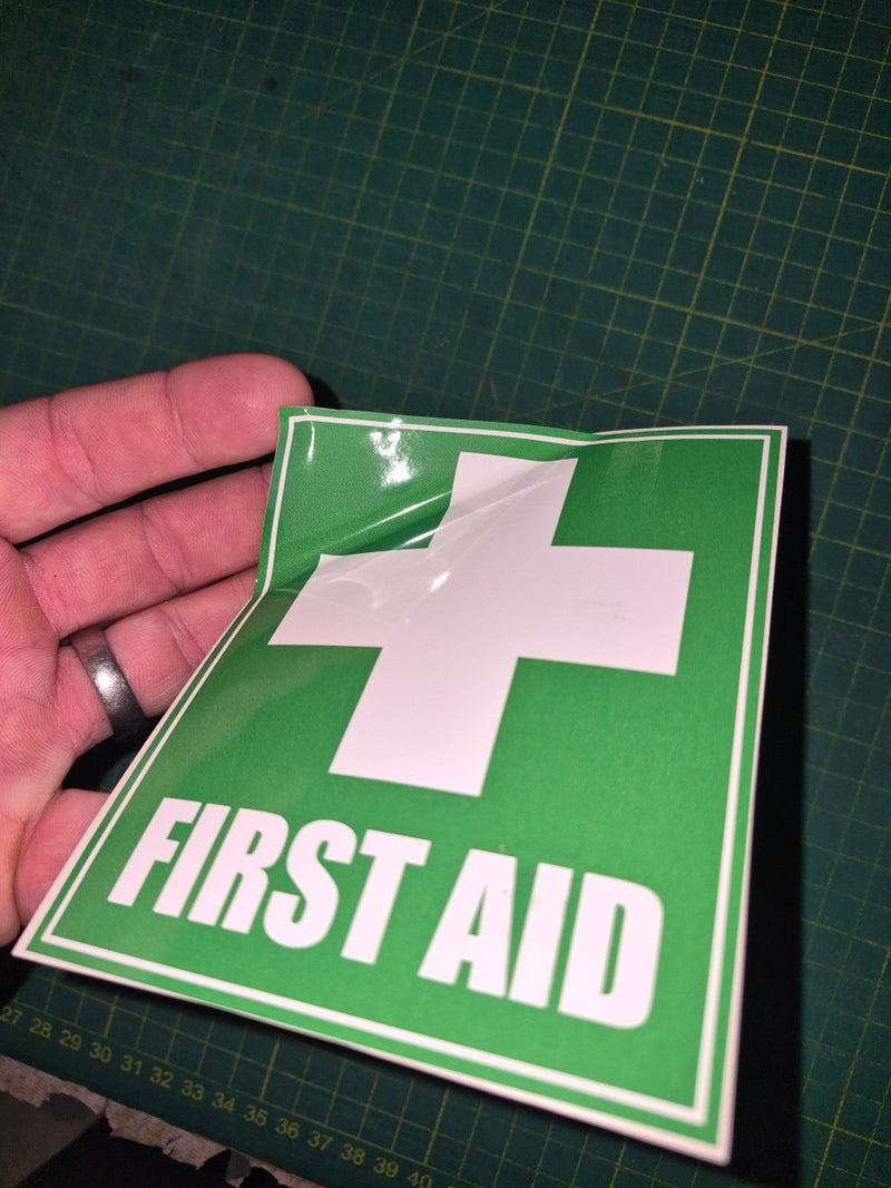 Green 'First Aid' sign with a white cross held by a hand on a grid background