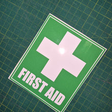 Load image into Gallery viewer, First aid sign with a white cross on a green background, placed on a grid surface.

