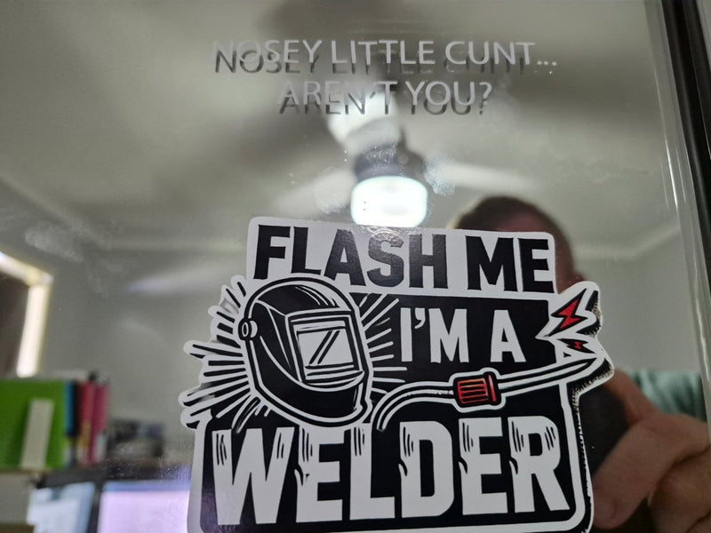 Sticker with text 'Flash me I'm a welder' held up to a mirror with offensive text.