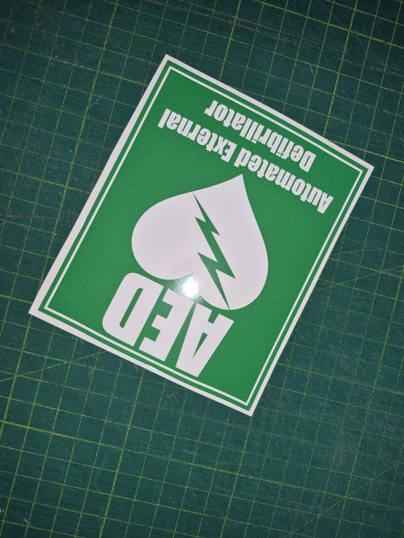 Green and white sticker with AED (Automated External Defibrillator) text and symbol on a grid background