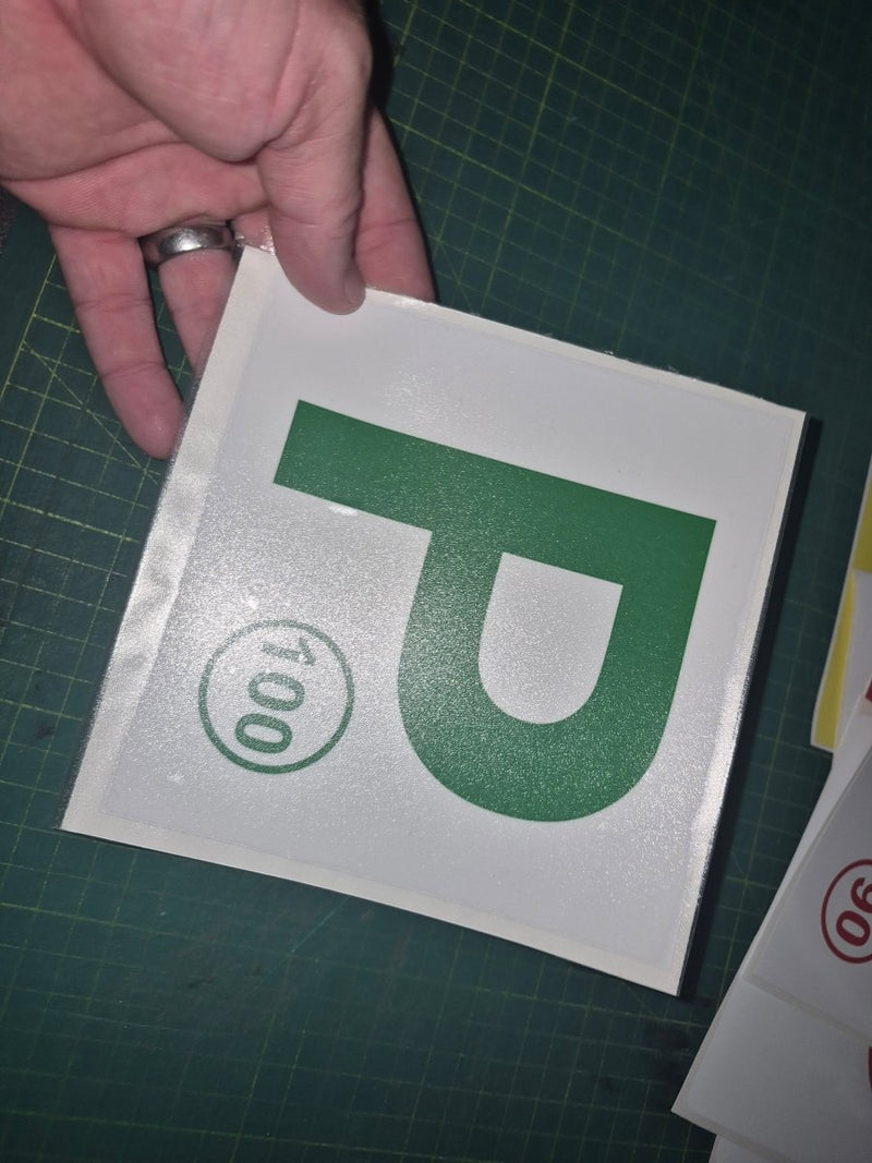 Hand holding a square decal with a green 'P' and number '100' on a green cutting mat.